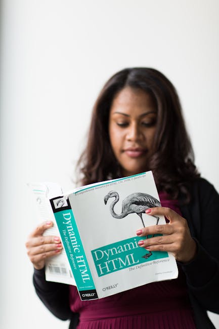 A woman with dark hair, wearing a black blazer over a purple top, is holding and reading a book titled 'Dynamic HTML' by O'Reilly, featuring a detailed black and white illustration of a flamingo on th